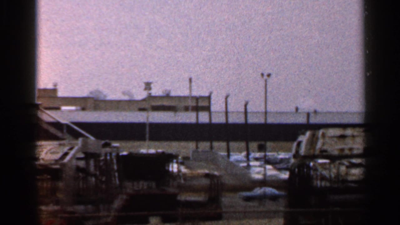 1966 Florida 8mm frame showing a fenced industrial or commercial lot with parked cars, low structures and a small distant tower.