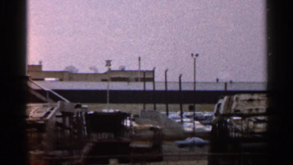 1966 Florida 8mm frame showing a fenced industrial or commercial lot with parked cars, low structures and a small distant tower.