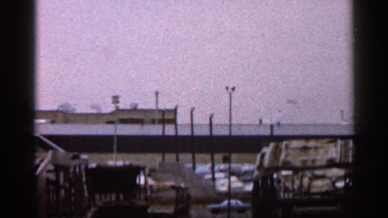 1966 Florida 8mm view across a fenced lot toward low buildings and parked vehicles, with dark home-movie framing at the edge.