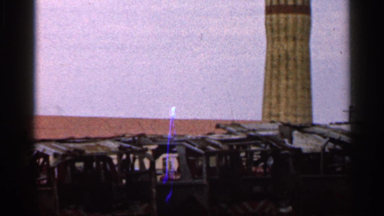 1966 Florida 8mm frame showing worn vehicles or trucks in a lot, a tall textured pillar, pale sky, and visible film-frame edges.
