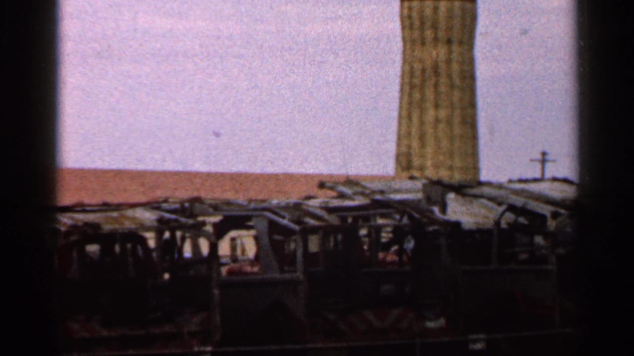1966 Florida 8mm frame of a junkyard-like vehicle lot, tall pillar, pale sky, and a small distant cross-shaped structure.
