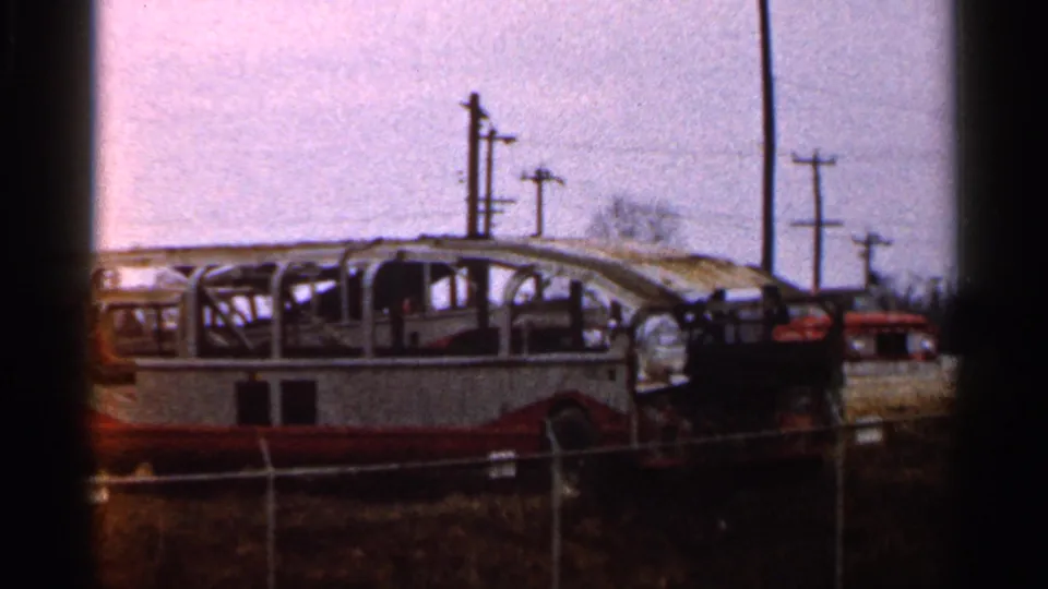 1966 home-movie frame showing a charred bus frame in a rural lot, seen through fencing under an overcast sky.