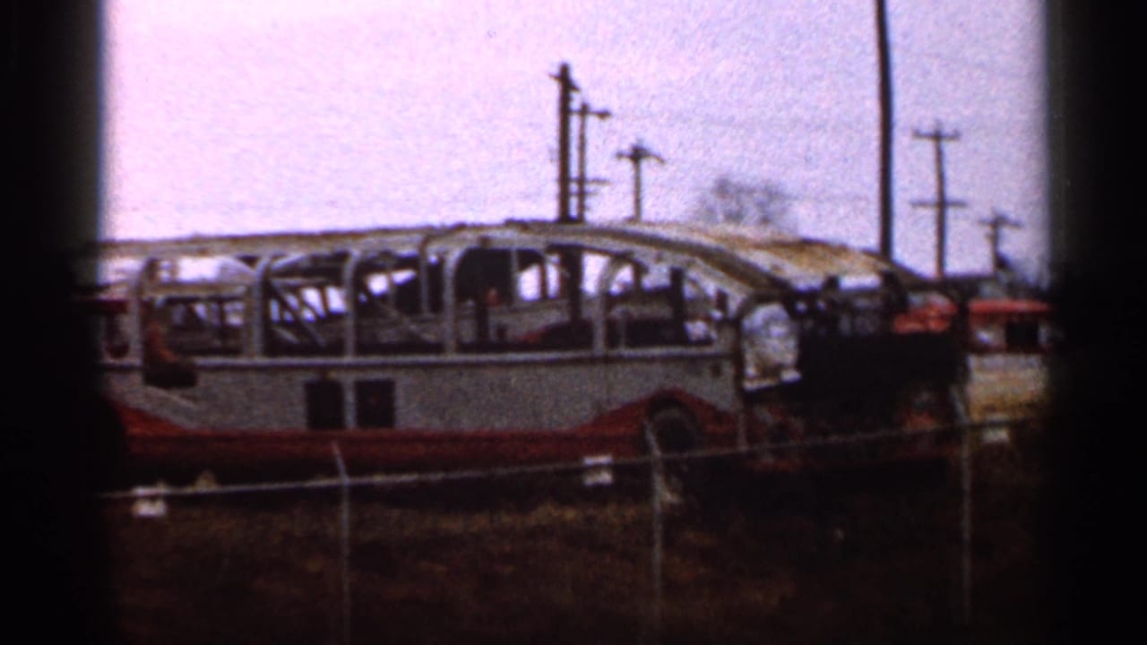 1966 8mm frame with the burned bus skeleton still centered, framed by fence lines and roadside utility poles.