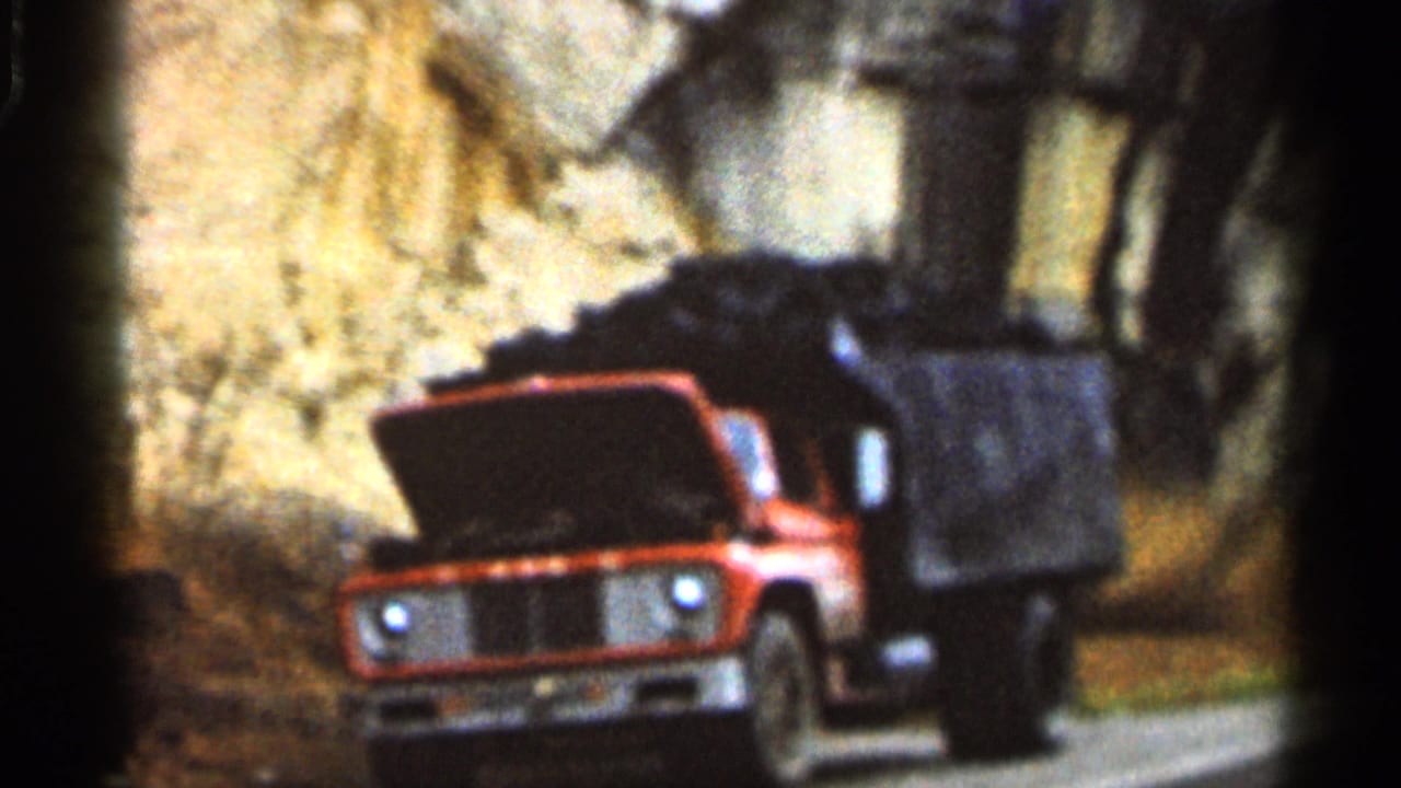 1966 Georgia home-movie frame showing a red-and-white dump truck with its hood open on a dirt road near a rocky roadside.