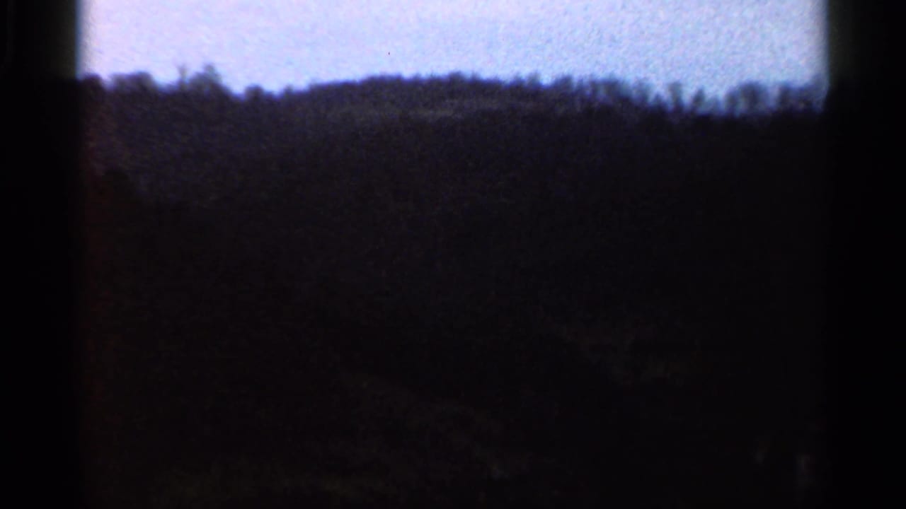 Grainy 1966 8mm view of rural hills and treetops under a cloudy sky, with dark exposure and soft detail.