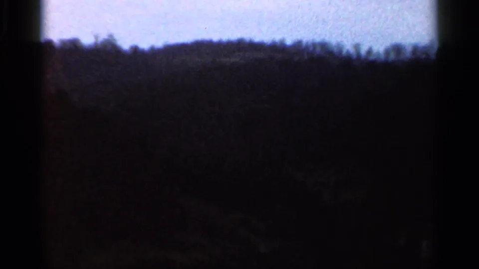 Grainy 1966 8mm view of rural hills and treetops under a cloudy sky, with dark exposure and soft detail.