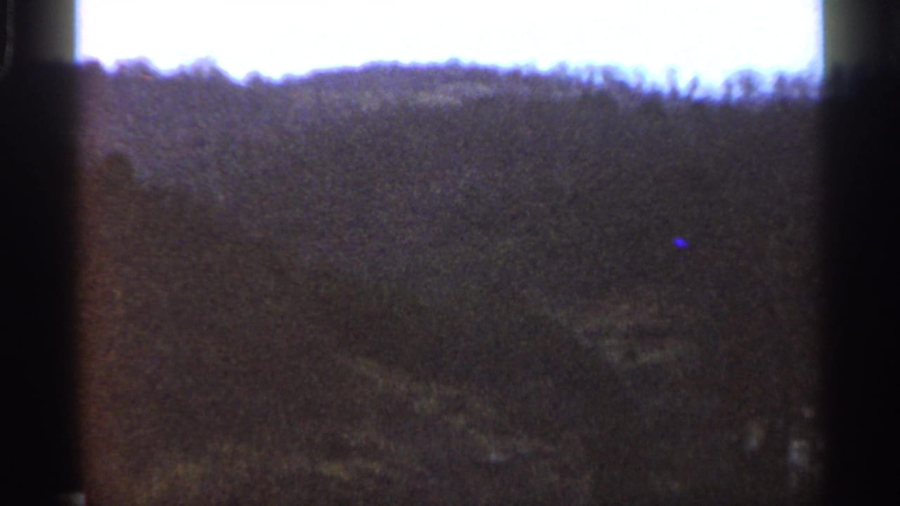1966 8mm landscape frame showing rolling Georgia hills, shrubs, and a bright cloudy sky in soft, grainy color.