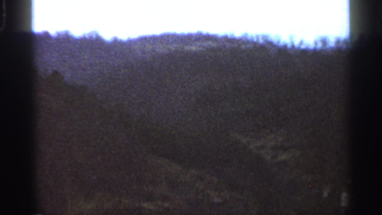 Dark wooded hillside below a bright overexposed sky, framed by 8mm film borders.