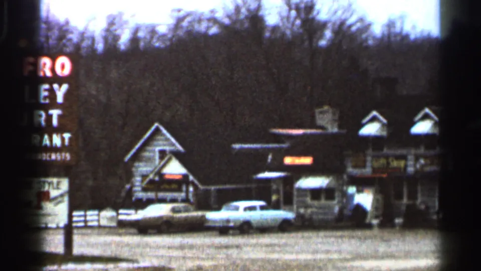 1966 Georgia home-movie frame showing rustic roadside shops, parked mid-century cars, a tall restaurant sign, gravel lot, and leafless trees.