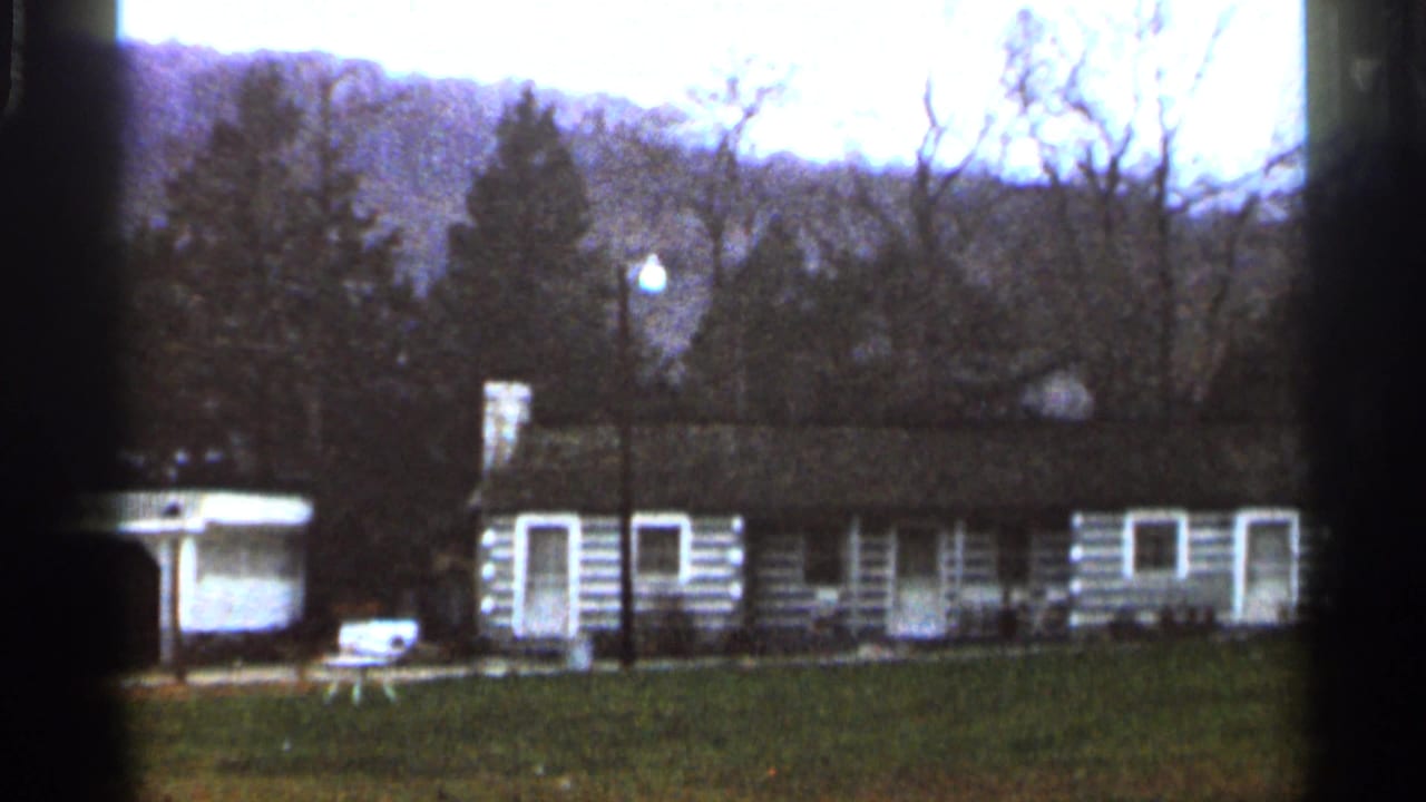 1966 8mm frame of a low rustic cabin structure with white trim, open lawn, bare trees, and rolling wooded hills.