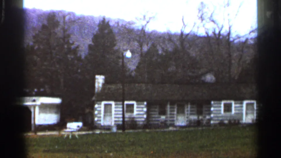 1966 8mm frame of a low rustic cabin structure with white trim, open lawn, bare trees, and rolling wooded hills.