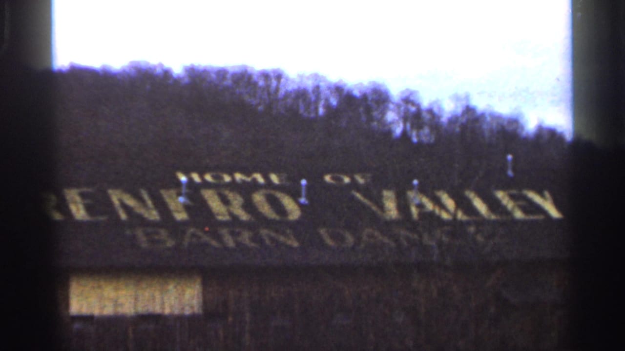 1966 8mm frame showing a weathered barn-like building with yellow Renfro Valley Barn Dance lettering and an overcast wooded hillside behind it.