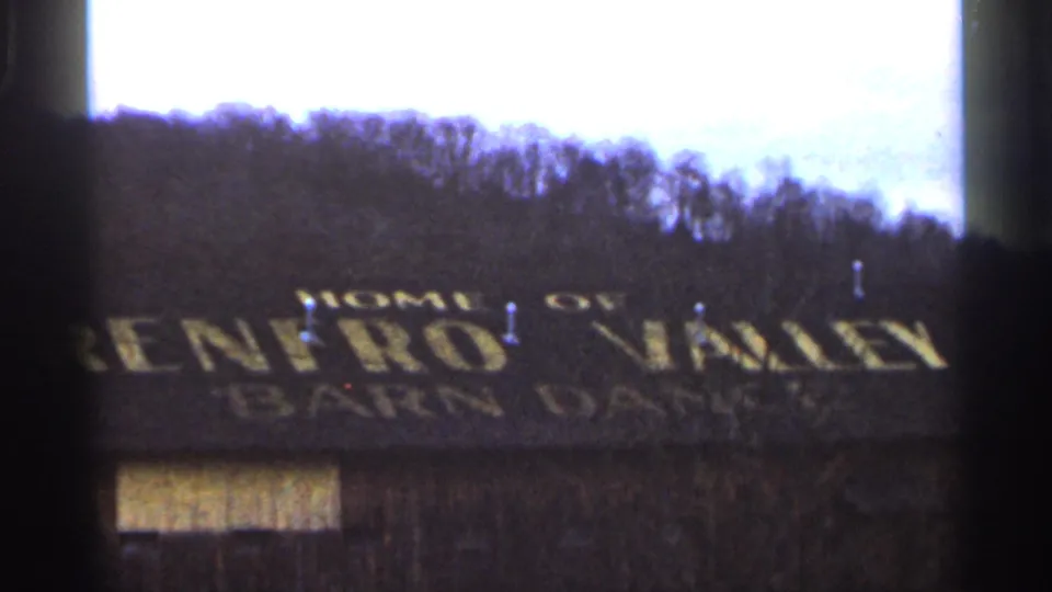 1966 8mm frame showing a weathered barn-like building with yellow Renfro Valley Barn Dance lettering and an overcast wooded hillside behind it.