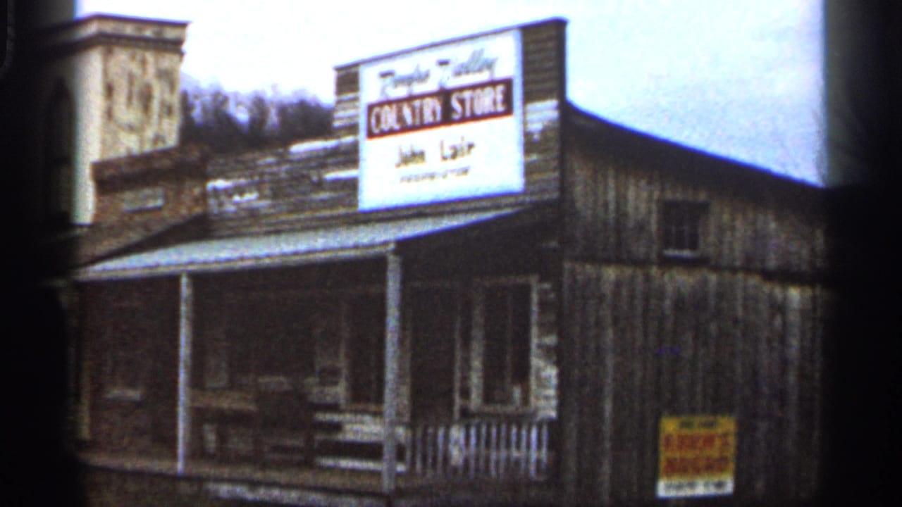 1966 home-movie frame of a rustic Country Store exterior with a prominent white sign, porch posts, wooden siding, and hillside in the background.