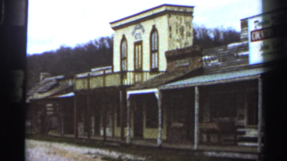 1966 8mm frame showing connected rustic storefronts with tall arched upper windows, covered porches, and a gravel roadside setting.
