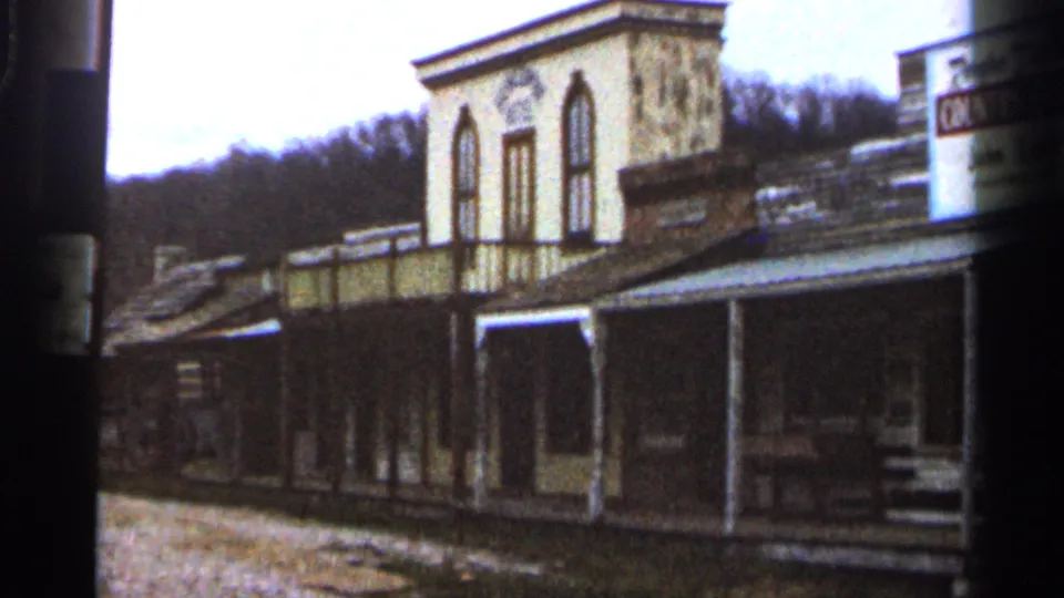 1966 home-movie frame of weathered wooden buildings with arched upper windows, porch posts, gravel roadway, and a partial Country Store sign at the edge.