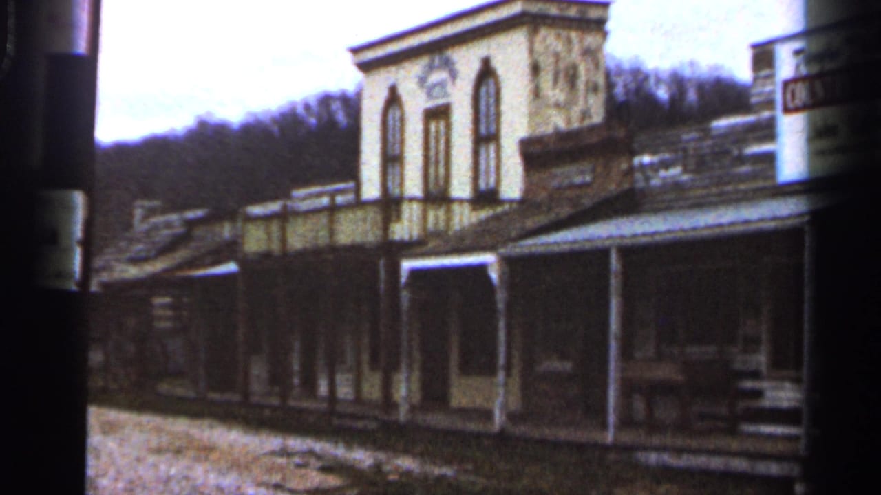 1966 Georgia home-movie frame showing weathered wooden storefronts, porch posts, and a two-story facade near wooded hills.