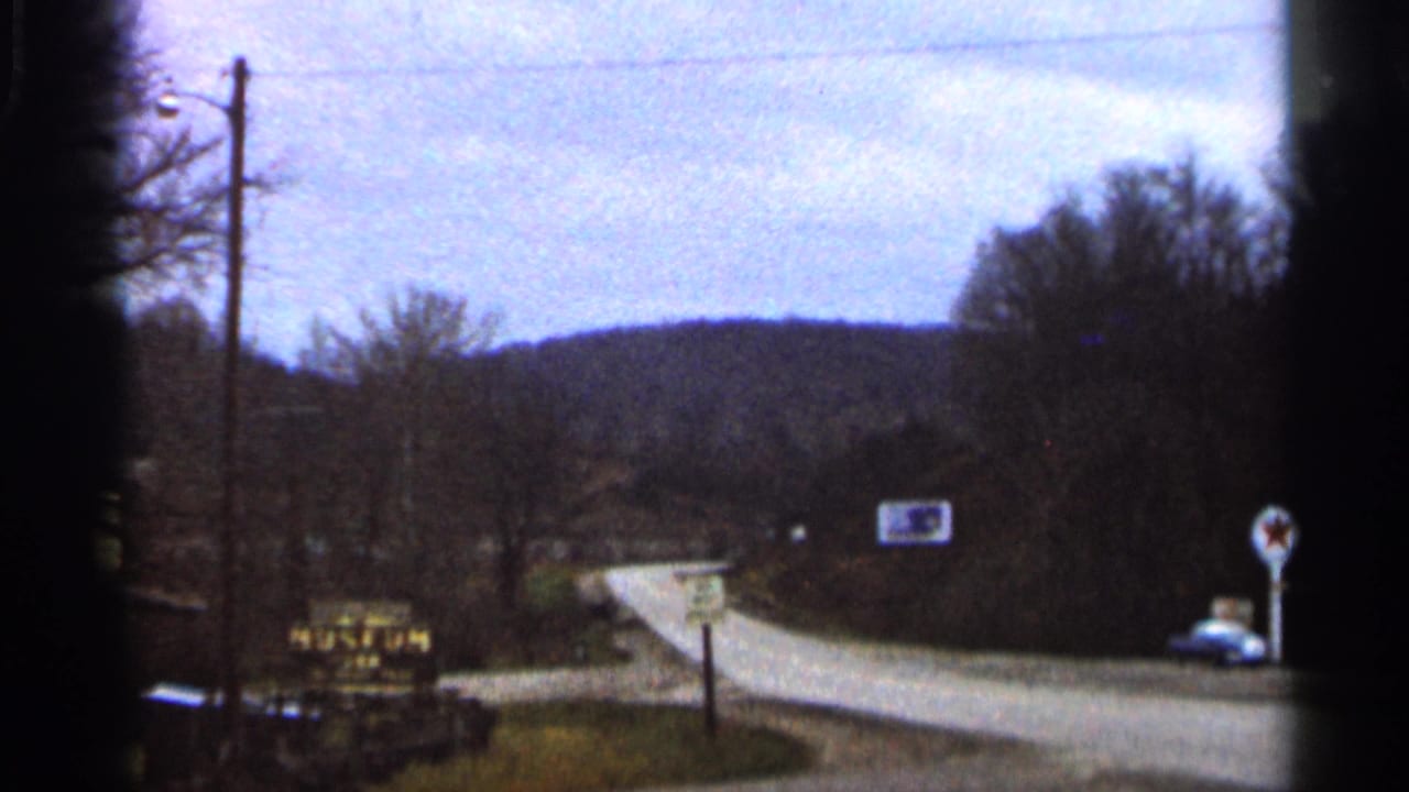 1966 Georgia road scene with wooded hills, a curving paved road, roadside signage, and a parked blue car near a service area.