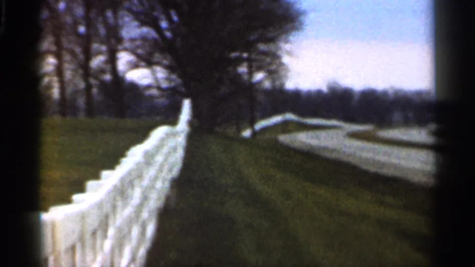 The pan reveals a winding road beyond the white fence, bordered by grass, bare trees, and a distant treeline in Georgia.
