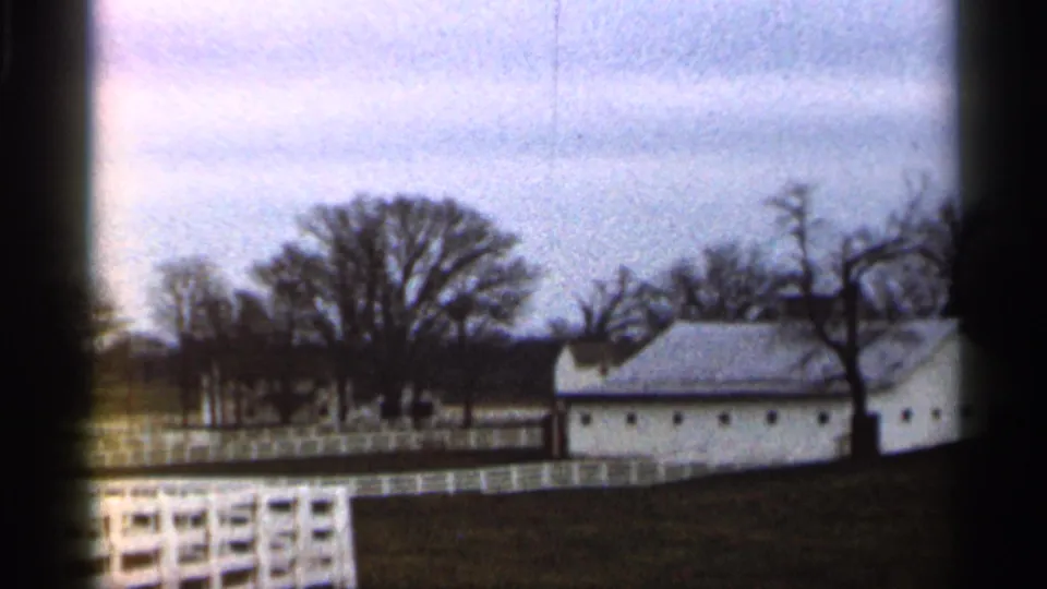 1966 Georgia rural view with a white farm building, white rail fencing, bare trees, and open grass in soft 8mm color.