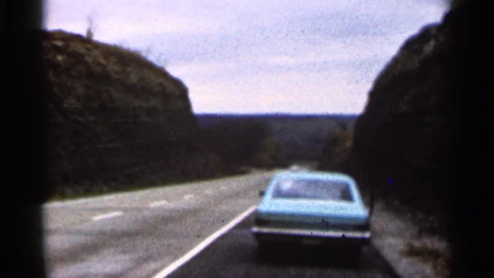 1966 Georgia road-trip view from a moving vehicle, following a light blue car along a narrow road between rocky cuts.