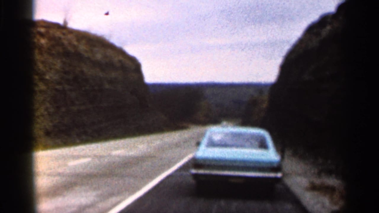 Blue car driving ahead on a two-lane road between rocky hills.