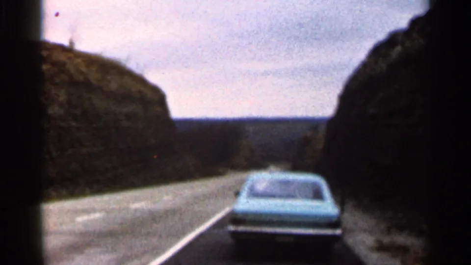 Blue car continues along a rural Georgia road in 1966, framed by dark rocky hills and muted 8mm color.