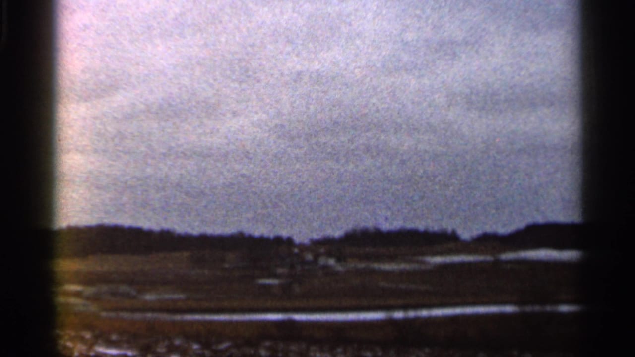Wide rural landscape with low hills, open ground, and bright reflective patches under cloudy sky.