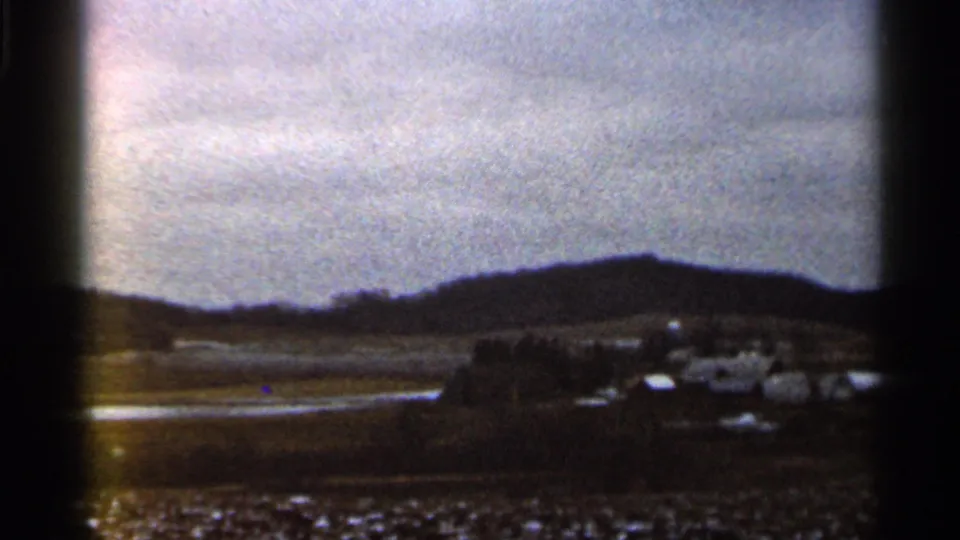 1966 Georgia 8mm frame showing low hills, scattered distant buildings, open ground, and reflective foreground areas under gray clouds.