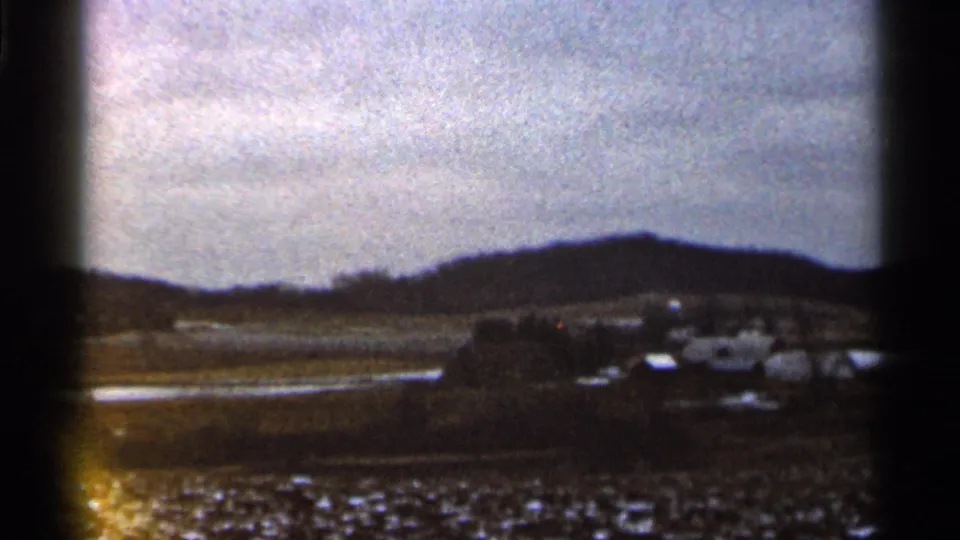 1966 Georgia home-movie frame with a dark hill ridge, small distant structures, open fields, and bright wet ground beneath heavy clouds.