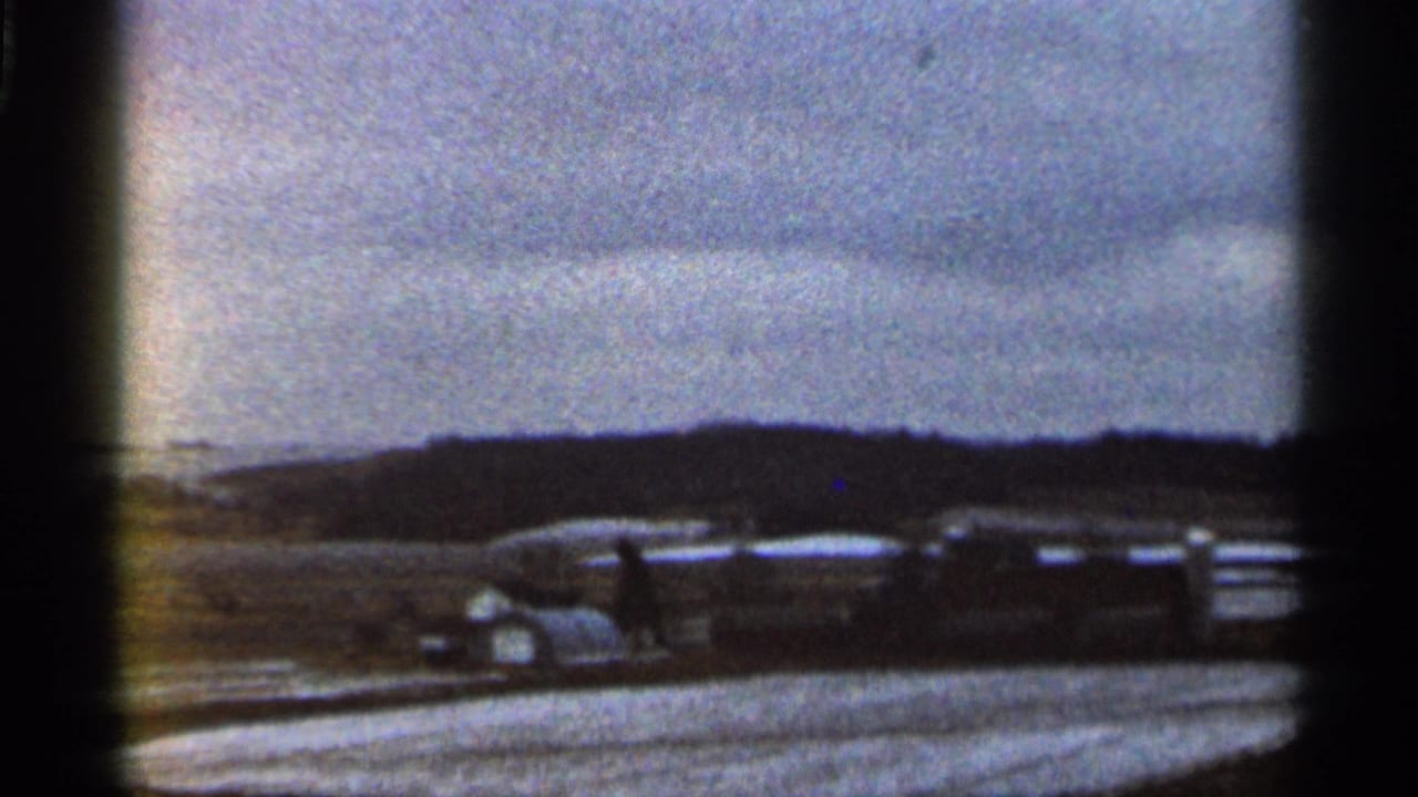 1966 Georgia 8mm frame showing vintage vehicles parked near open ground with low hills and a cloudy sky.