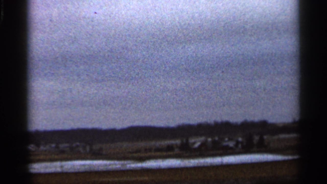 1966 8mm rural Georgia view with patchy snow, distant low buildings, bare horizon trees, and dark film-frame edges.