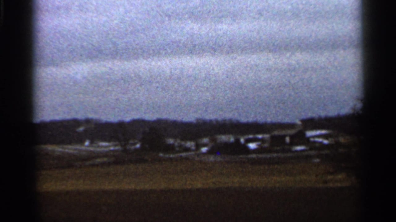 Distant farm buildings across an open brown field below layered clouds.