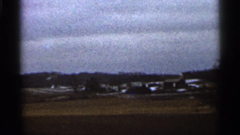 Rural Georgia winter landscape on 8mm film, with open field, low buildings, bare trees, and a dark horizon beneath cloud cover.