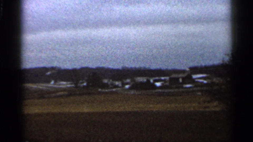 1966 home-movie frame of a rural Georgia field, showing snow patches, distant structures, bare trees, and muted 8mm color.