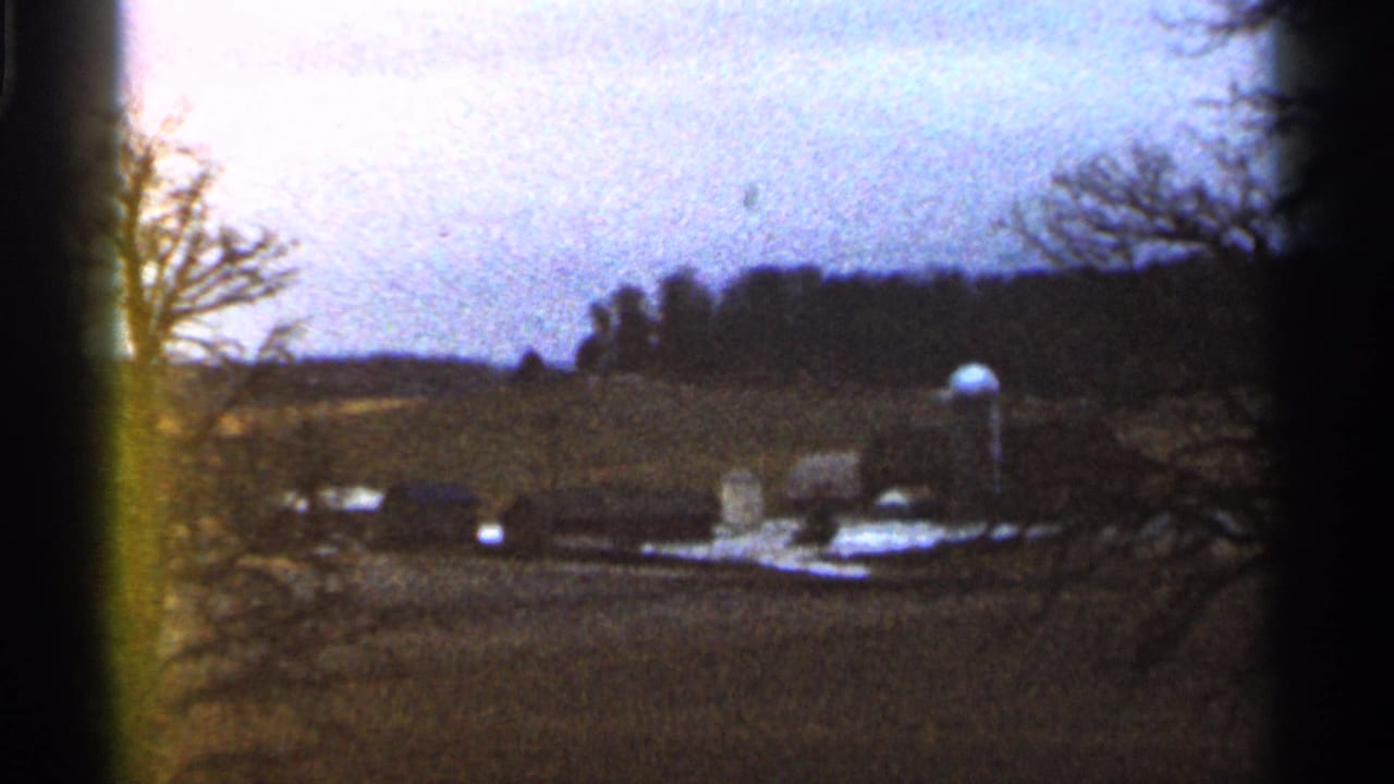 Distant rural field with bare trees, farm buildings, and a small water tank under a pale sky.