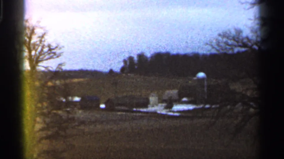 1966 Georgia home-movie frame with a distant water tank and low rural buildings seen across fields through soft 8mm focus.