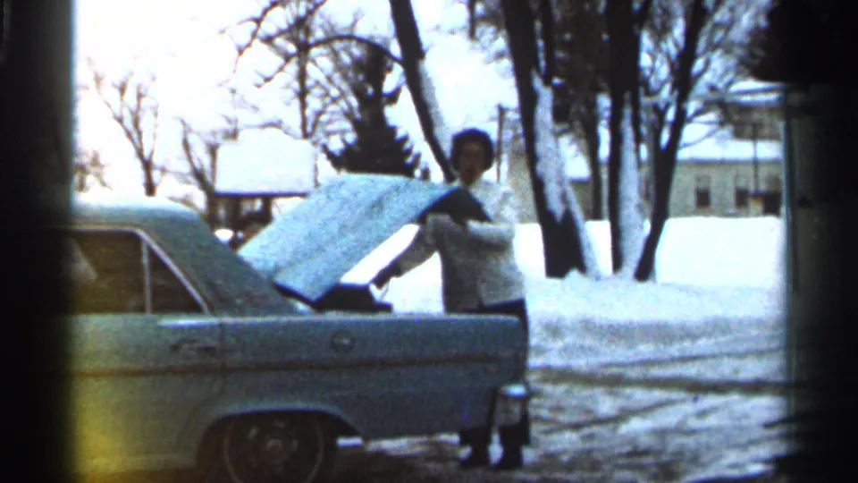 In the 1966 Georgia home-movie footage, a woman stands near a light blue car with the hood open, surrounded by snow and bare trees.