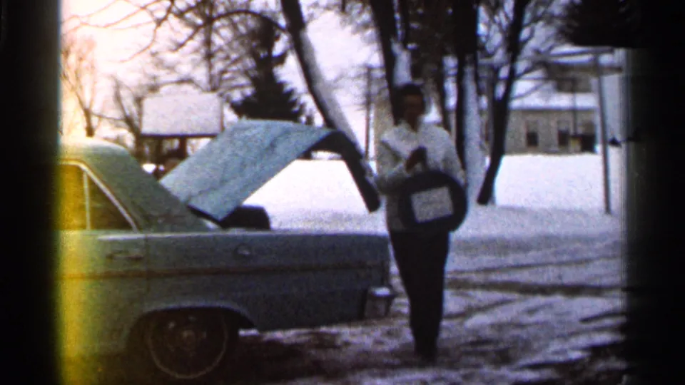 Woman in winter clothing holds a round dark car part or cover beside the light blue car, with snow, trees, and a house behind her.