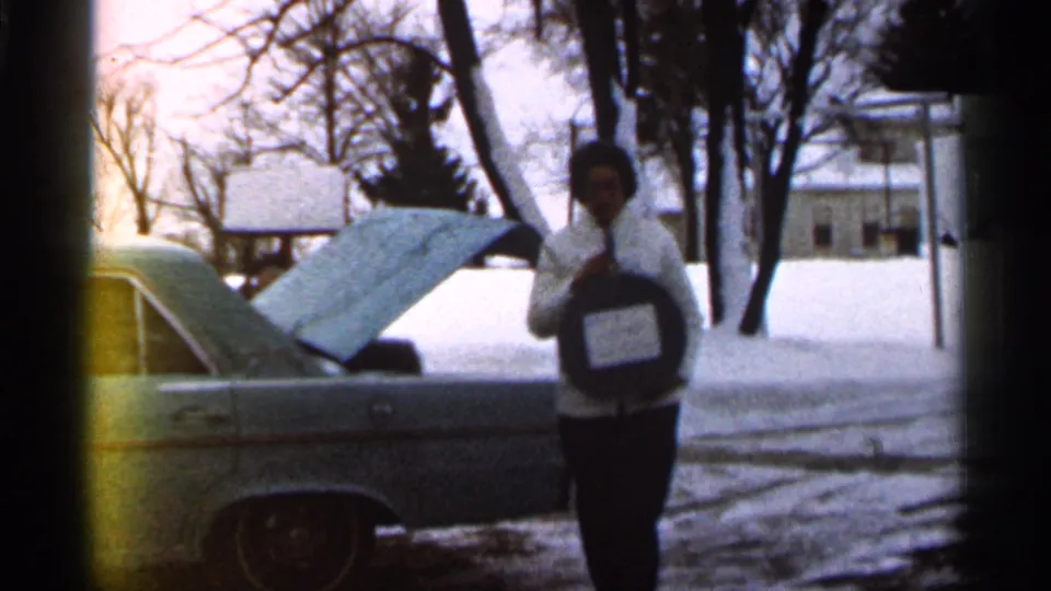 The woman faces the camera while holding a round dark object beside the open-hood car in a snowy Georgia yard, consistent with 1966 home-movie metadata.