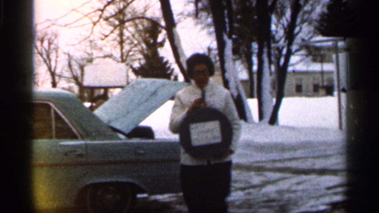In 1966 Georgia home-movie footage, a woman in glasses holds a round case beside a car with an open hood in a snowy residential yard.