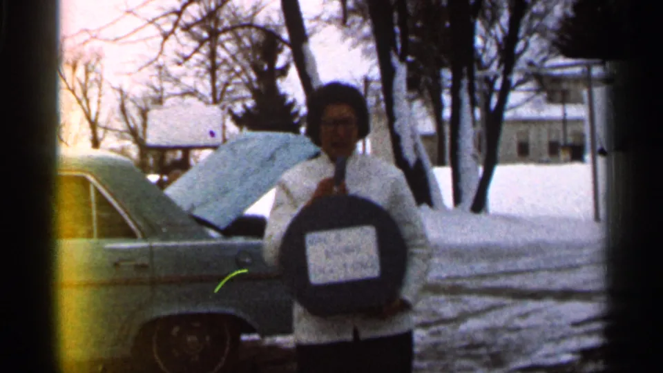 The woman stands closer to camera, holding a labeled round case while a vintage car and snowy yard remain visible behind her.