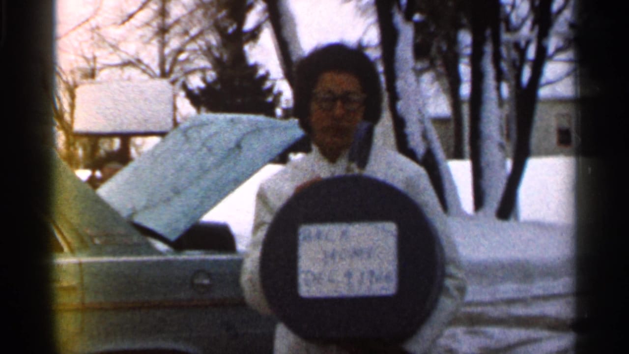 In 1966 Georgia home-movie footage, a woman in a white coat and glasses poses with a “BACK HOME” sign in a snowy residential yard.
