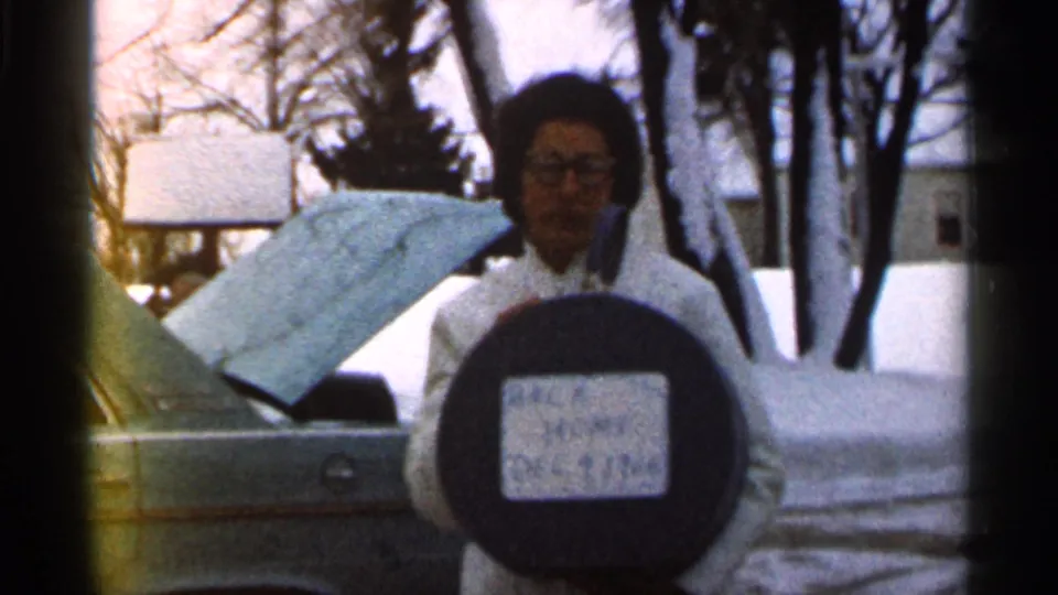 In 1966 Georgia home-movie footage, a woman in a white coat and glasses poses with a “BACK HOME” sign in a snowy residential yard.
