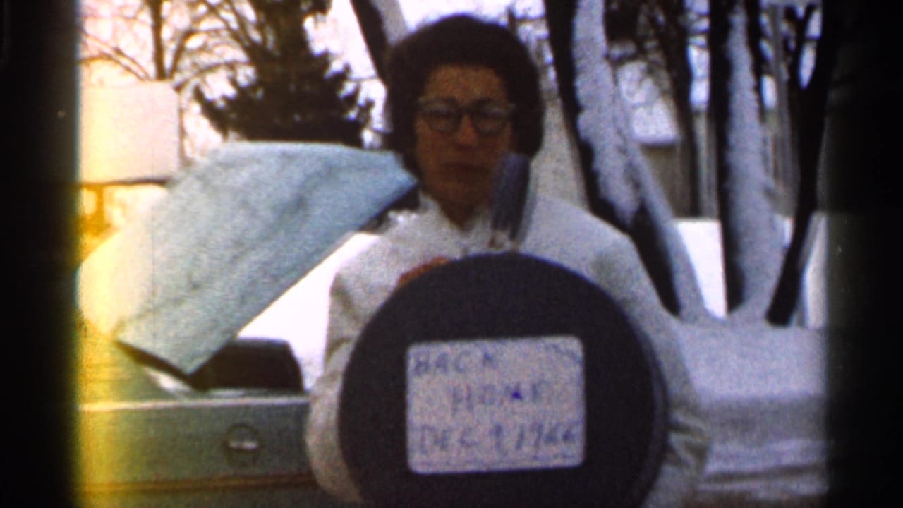 Woman faces the camera holding a round sign, with snowbanks, trees, and a car behind her.
