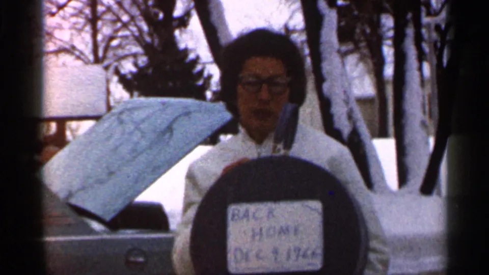 A softly focused 8mm frame shows the woman with the circular sign beside an open car hood, snow, trees, and residential buildings.