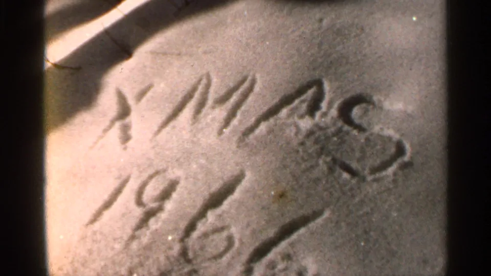 Close-up home-movie frame of “XMAS 1966” etched into a pale snowy or frosted surface, with soft grain and warm exposure.