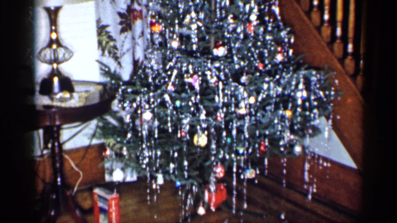 1955 New York home-movie frame showing a tinsel-covered Christmas tree near a table lamp, floral curtains, and wood trim.
