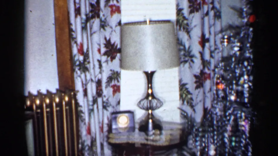 1955 New York living room detail with a radiator, table lamp, small clock, floral curtains, blinds, and the edge of a tinsel tree.