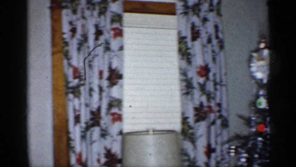 1955 New York home interior showing floral curtains, closed blinds, a lamp shade below, and part of a decorated Christmas tree at right.